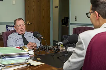 Man in a gray sweater sits at a desk with a stack of papers, speaking into multiple microphones during an interview in a small studio.