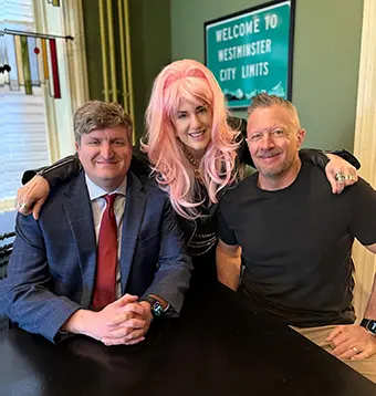 Three people sit at a table smiling; the middle person wears a pink wig and flanks two men in casual/business attire.