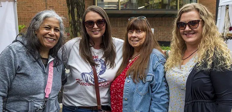 Group of four women smiling for the camera outdoors, wearing casual jackets and sunglasses.