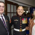 Three smiling adults at a formal event: a Marine in dress uniform with medals flanked by a man in a suit and a woman in a tan jacket nearby.