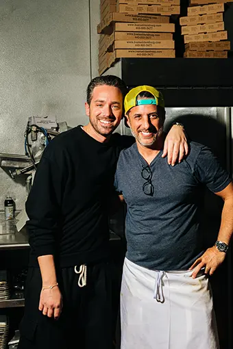 Louie Vassilaras and Yianni Livaditis - Carroll News | Carroll Publications | Carroll County, MD Two men in a kitchen pose for a photo; one wears a black hoodie, the other a gray shirt with a yellow cap and apron, smiling with arms around each other.