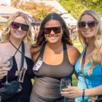 Three women wearing sunglasses pose together at an outdoor festival, each holding a wine glass.