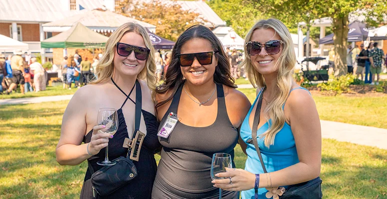 Three women wearing sunglasses pose together at an outdoor festival, each holding a wine glass.