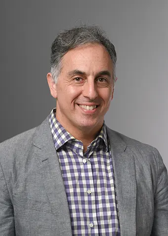 Smiling middle-aged man in a gray blazer and purple plaid shirt against a neutral gray background.