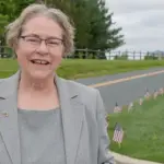 Smiling older woman in a gray blazer stands beside a road with a row of small American flags on the grass behind her.