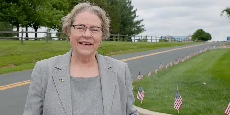 Smiling older woman in a gray blazer stands beside a road with a row of small American flags on the grass behind her.