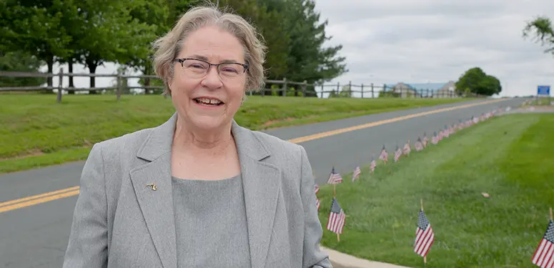 Smiling older woman in a gray blazer stands beside a road with a row of small American flags on the grass behind her.