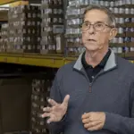 Older man wearing glasses speaks and gesticulates in a warehouse aisle with shelves of boxed goods behind him, suggesting a briefing or tour.