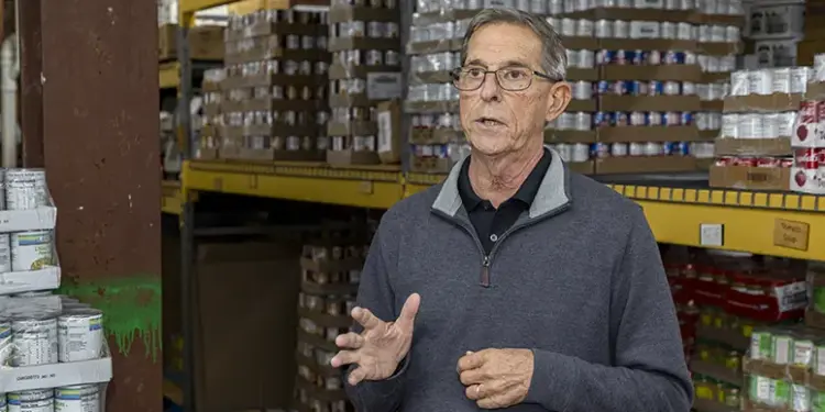 Steve Lambertson Talking - Carroll News | Carroll Publications | Carroll County, MD Older man wearing glasses speaks and gesticulates in a warehouse aisle with shelves of boxed goods behind him, suggesting a briefing or tour.