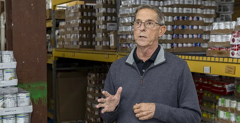 Older man wearing glasses speaks and gesticulates in a warehouse aisle with shelves of boxed goods behind him, suggesting a briefing or tour.