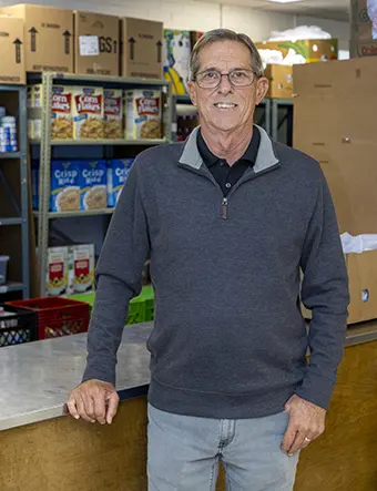 Steve Lambertson - Carroll News | Carroll Publications | Carroll County, MD Smiling older man with glasses behind a counter in a food pantry; shelves of cereal and cardboard boxes in the background.