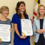 Three women stand indoors, two in blue-toned dresses holding framed certificates and smiling as the third woman in a light blue blazer stands beside them.