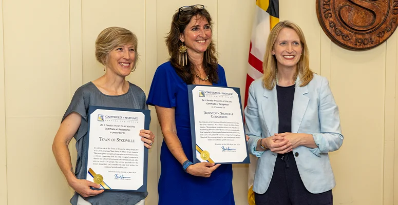 Three women stand indoors, two in blue-toned dresses holding framed certificates and smiling as the third woman in a light blue blazer stands beside them.