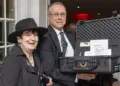 Two smiling adults pose at an event, with a man holding an open hard-shell equipment case containing papers and items for auction or raffle in a banquet setting.