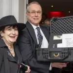 Two smiling adults pose at an event, with a man holding an open hard-shell equipment case containing papers and items for auction or raffle in a banquet setting.
