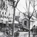 Historic city street with horse-drawn carriages, vendors, and pedestrians along a row of shops on a tree-lined avenue.