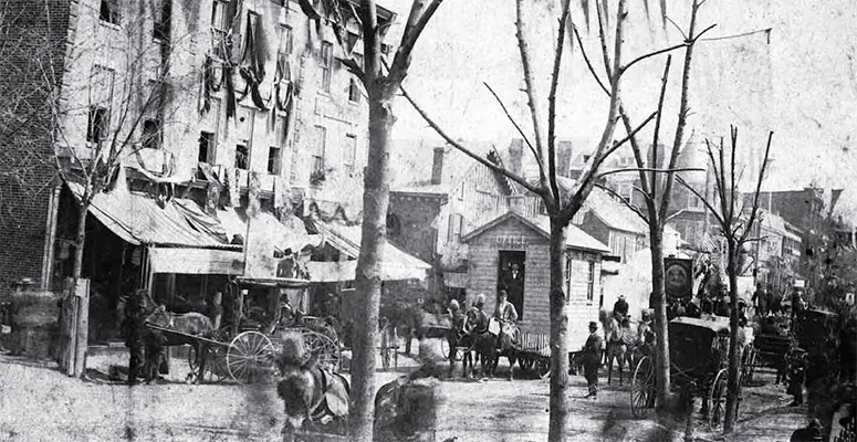 Historic city street with horse-drawn carriages, vendors, and pedestrians along a row of shops on a tree-lined avenue.
