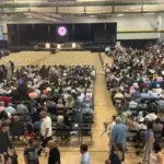 Large school gym packed with hundreds of attendees seated and facing a stage with screens on either side.