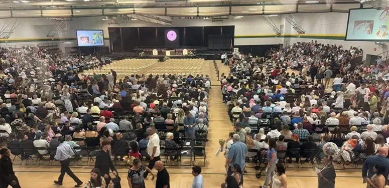 Large school gym packed with hundreds of attendees seated and facing a stage with screens on either side.