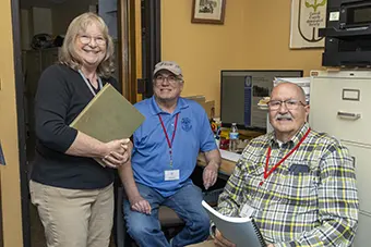 Three colleagues in an office: a smiling woman stands with a folder while two men sit with lanyards nearby.