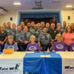Group photo of sheriff's deputies and volunteers at a community event behind a table with The Shepherd's Staff banners in a gym setting
