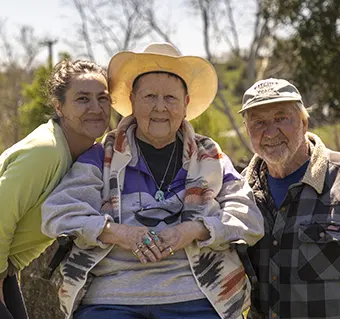 Three people outdoors posing for a photo, with an elderly woman in the center wearing a wide-brim hat, flanked by a smiling couple on each side.